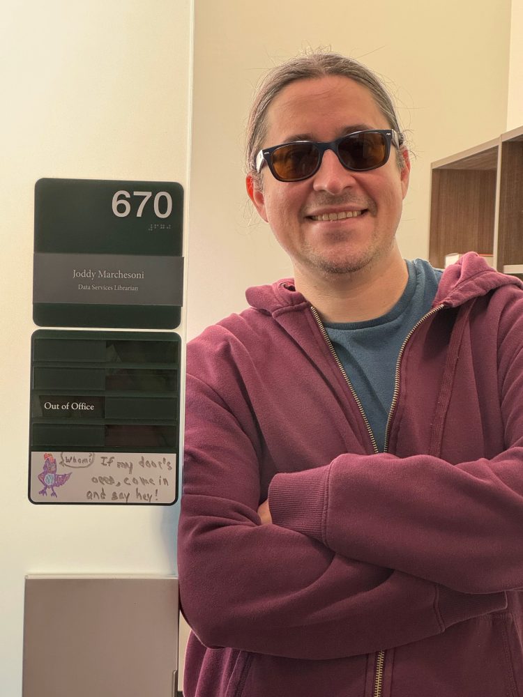 A librarian in a red jacket and sunglasses smiles with their arms crossed.