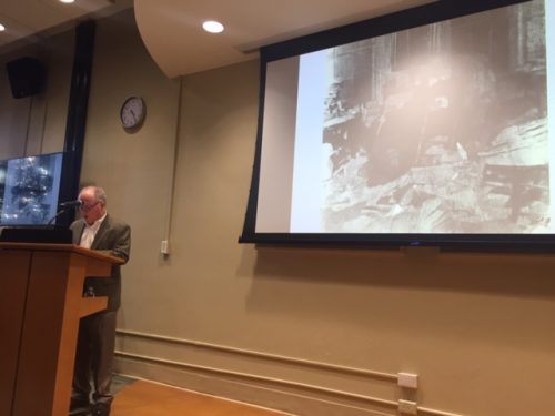 man stands at podium in front of an image of Walt Whitman