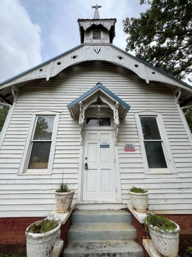 A weathered white-painted wood and brick church, with blue accents and potted plants on a set of front steps.