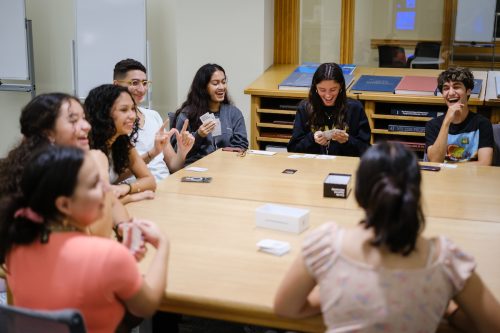 A group of eight students sit around a table, playing Cards Against Humanity